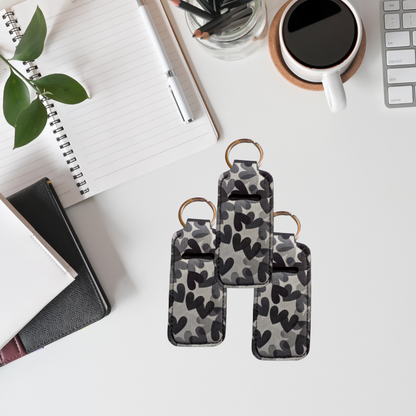 Three camouflage-patterned keychains on a desk with a notebook, plant, and coffee cup in the background.