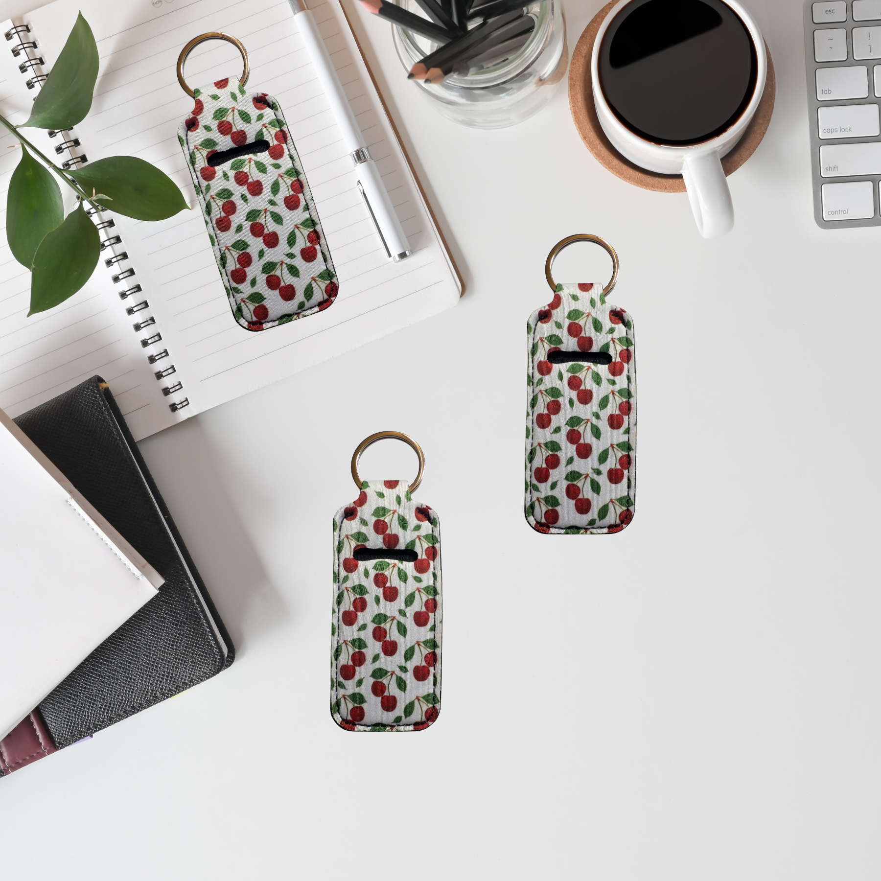 Three cherry-patterned keychain pouches on a desk with a coffee cup, plant, and office supplies.