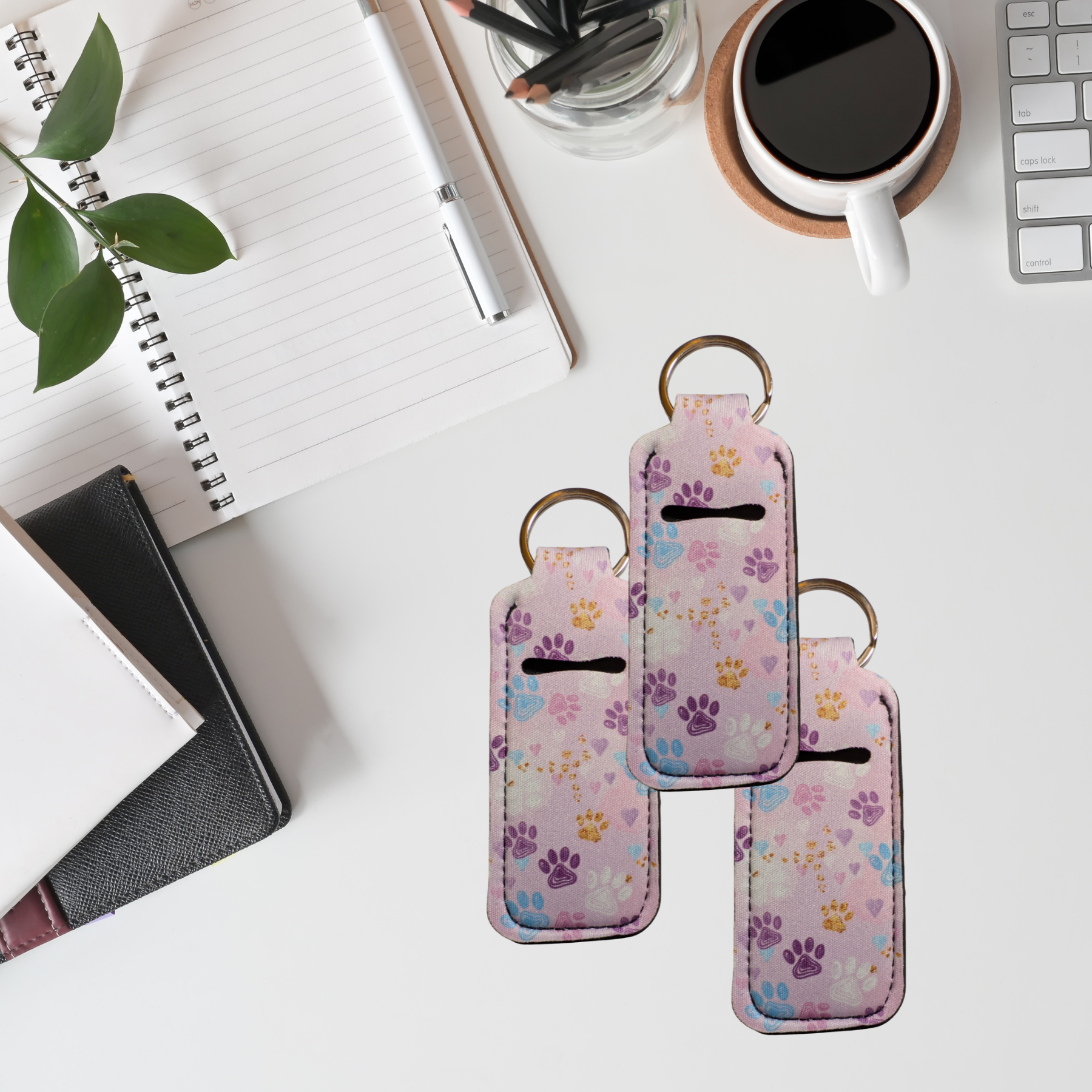Three pink phone cases with paw prints on a desk with a notebook, coffee, and plant.