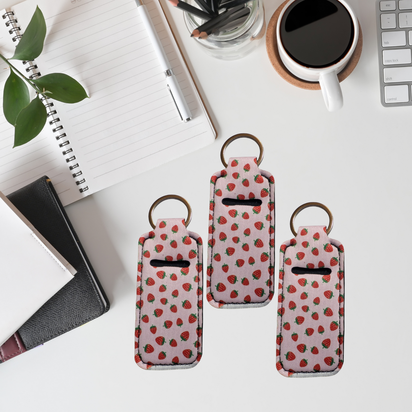 Three strawberry-patterned keychains on a desk with a notebook, plant, and coffee cup in the background.
