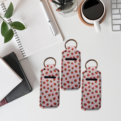 Three strawberry-patterned keychains on a desk with a notebook, plant, and coffee cup in the background.