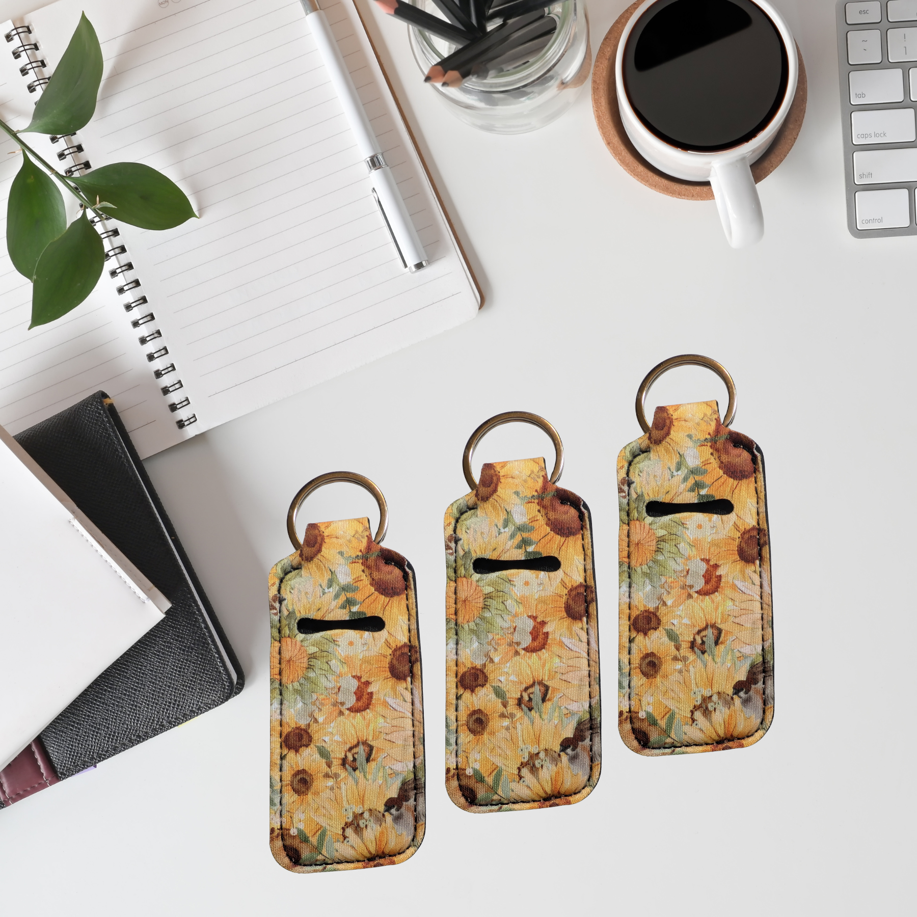 Three floral keychains on a desk with a notebook, coffee cup, and plant.